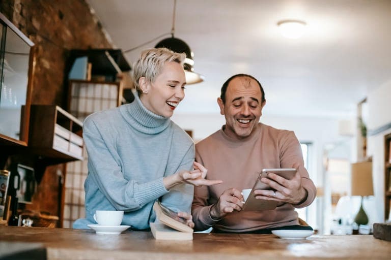 Two adults smile and interact while looking at a tablet at a cozy café setting.