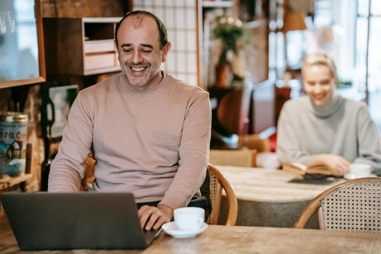 A man smiles while working on a laptop at a café table, with a woman reading in the background.