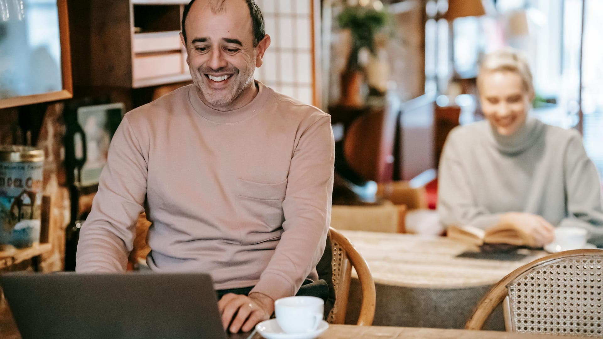 A man smiles while working on a laptop at a café table, with a woman reading in the background.