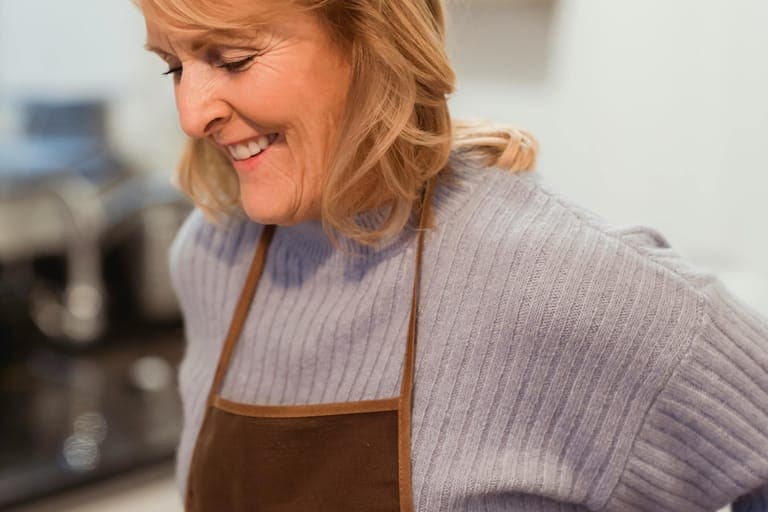 A smiling woman in a blue sweater and brown apron stands in a kitchen.